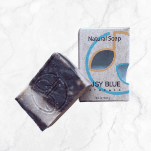 A Purely Unscented Bar Soap with a swirl pattern rests beside its box, both featuring a stylized logo and "Natural Soap" with "Daisy Blue Naturals," arranged on a white marble surface.