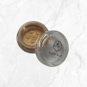 A clear-lidded round container labeled "Daisy Blue Naturals" holds loose, shimmery gold Flawless Glow Blush with sifter holes visible in the open lid, resting on a white marble surface.