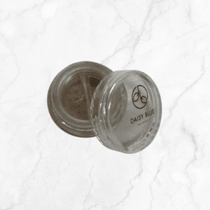 A small, round clear container with a sifter lid holds Flawless Glow Blush. The "DAISY BLUE NATURALS" label and logo appear on the lid, as the blush sits elegantly on a white marble surface.