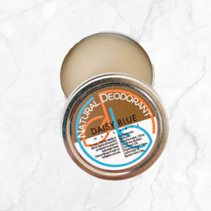 A round tin of Natural Deodorant Cream sits on a white marble surface, its lid partially open to reveal the solid balm inside. The colorful label lists ingredients in orange, brown, and blue.