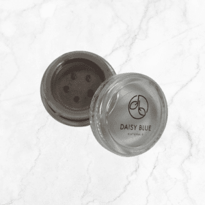 A small, clear container of Flawless Glow Blush with a sifter lid—partially open to show the powder inside—sits on a white marble surface. The lid features the DAISY BLUE NATURALS logo and branding.