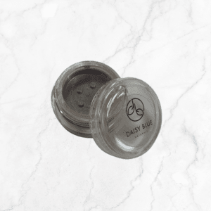 A small, round, clear plastic container with a sifter lid and the "Daisy Blue Naturals" logo holds Flawless Glow Blush loose powder. The container sits on a white marble surface with its lid slightly open.