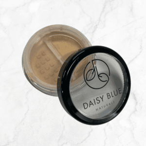 An open container of Flawless Glow Blush loose powder makeup with a black lid featuring the "Daisy Blue Naturals" logo rests on a white marble surface.