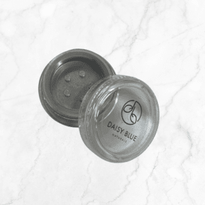 An open clear plastic container of Flawless Glow Blush with a sifter lid displays its contents on a white marble surface. "Daisy Blue Naturals" is labeled on the packaging.