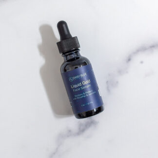 A dark glass dropper bottle of Liquid Gold Face Serum rests on a white marble surface, featuring a black cap and a blue label with crisp white text.