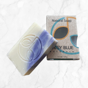 A rectangular Purely Unscented Bar Soap with cream and purple swirls rests next to its "Natural Soap" and "Daisy Blue Naturals" labeled box on a light marble background.
