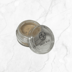 A small glass jar labeled "DAISY BLUE NATURALS" holds Flawless Glow Blush, featuring light beige powder, displayed elegantly on a white marble surface.