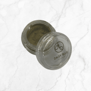 An open round container of Flawless Glow Blush makeup powder with a clear lid, labeled "Daisy Blue Naturals," sits on a white marble surface. The shimmery gold blush is visible inside the packaging.
