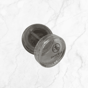 An open, small round clear plastic container labeled “Daisy Blue” sits on a white marble surface, showing the Flawless Glow Blush—a brownish cosmetic product—inside its transparent lid.