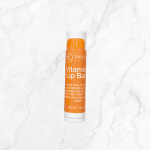 An orange tube of Daisy Vitamin Lip Balm rests on a white marble surface. The cruelty-free, flavor-free, petroleum-free formula is ideal to use with Flawless Glow Blush. Net weight: 0.2 oz (5.9 ml).