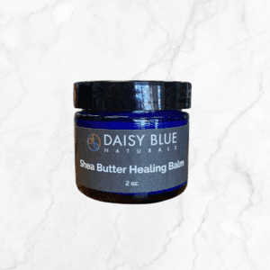 A blue jar of Shea Butter Healing Balm with a black lid and minimalist design sits on a white marble surface. This balm is perfect for soothing and nourishing your skin.