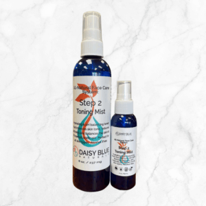 Two blue bottles of Daisy Blue Naturals Facial Toning Mist, labeled "Step 2 Toning Mist" and featuring white caps in large and small sizes, are displayed on a light marble background.