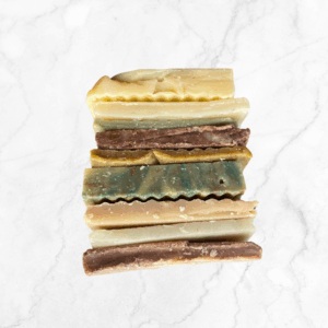 The Soap Stack: Odds & Ends displays five assorted handmade soaps with wavy edges in yellow, brown, blue-green, and beige hues, arranged vertically on a white marble background.
