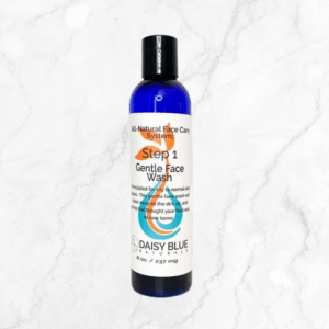A blue bottle of Gentle Face Wash stands upright on a white marble surface. Its white label features orange and black text highlighting the product as an all-natural formula.