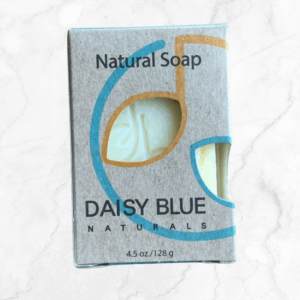 A rectangular box of Orange Blossom Body Bar Soap with a cut-out displays the light yellow soap inside. The packaging features blue and gold curved lines and is placed on a white marble surface.