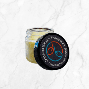 An open small glass jar of Copaiba Balsam Therapeutic Salve reveals the creamy salve inside. The black lid features the colorful "Daisy Blue Naturals" logo, set on a white marble background.