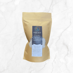 A brown resealable pouch labeled "Copaiba Balsam Bath Salts" sits on a white marble surface, featuring a clear window that reveals the white bath salts inside.