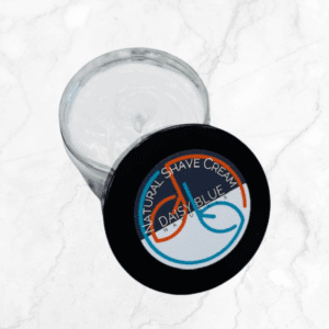 An open Natural Shave Cream jar with a black lid labeled "Natural Shave Cream" rests on white marble. The smooth, white cream inside offers a gentle, effective shaving experience.