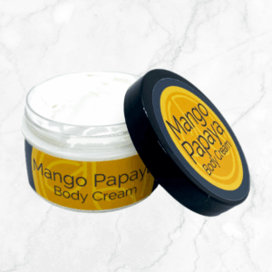 An open jar of Mango Papaya Body Cream with a yellow and orange label rests on a light marble surface, its lid—showing the product name—tilted nearby to reveal the creamy white, fruit-infused formula inside.
