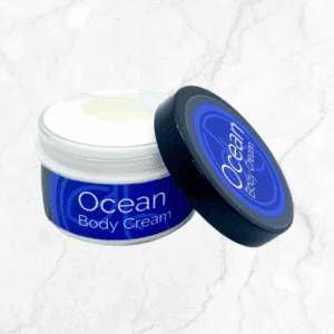An open jar of Ocean Body Cream with a blue label and matching lid sits on white marble. The lid rests beside the jar, revealing the luxurious cream inside.