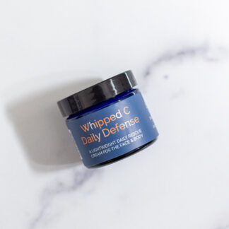 A blue jar labeled “Whipped C Daily Defense” with a black lid sits on a white marble surface, casting a soft shadow. Whipped C Daily Defense is formulated to protect and rescue your face and body daily.