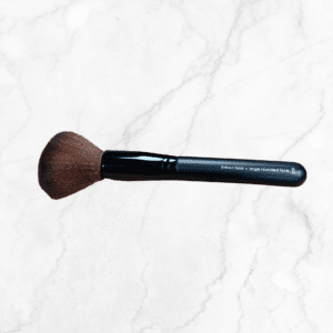 A Full Face Brush with soft brown bristles and a black handle, labeled "brown faux - large rounded face," rests on a white marble surface.