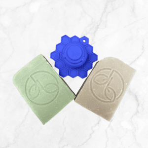 A green and a beige Face Cleansing Bar with leaf logos rest side by side on white marble, topped by a blue, honeycomb-patterned ScrubBEE silicone holder. Product: Face Cleansing Bar with ScrubBEE.