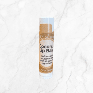A tube of Coconut Lip Balm with a beige label stands upright on a white marble surface. The label reads: "Softens and nourishes lips with all-natural ingredients.