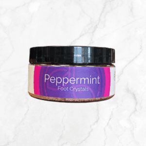 A round jar labeled "Peppermint Foot Crystals, NEW SIZE" with a black lid rests on a purple and white background atop white marble—ideal for refreshing foot care with the soothing scent of peppermint.