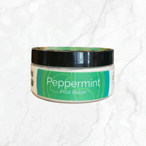 A round container of Peppermint Foot Butter - NEW SIZE with a green label rests on white marble, its name prominent on the jar, offering soothing relief for tired feet.