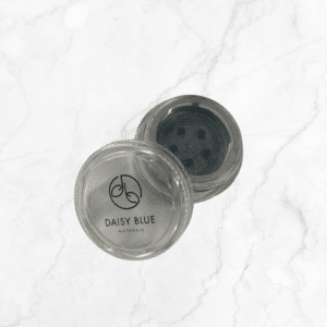 An open, round, clear container labeled "DAISY BLUE NATURALS" on the lid reveals a sifter insert and displays Charcoal Mist Eyeshadow on a white marble surface.