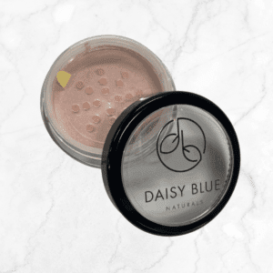 An open container of Dusty Rose Blush loose powder, labeled "DAISY BLUE NATURALS," rests on a white marble surface. This shimmery light pink blush is perfect for adding a radiant glow to your look.