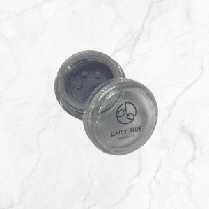 An open small, round, clear container of Charcoal Mist Eyeshadow with a sifter lid sits on a white marble surface. The product name and brand logo are visible on the lid.